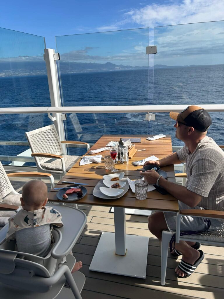 Papa und Baby beim Essen im Außenbereich des X-View und Blick aufs Meer bei einer Kreuzfahrt mit der Mein Schiff Relax